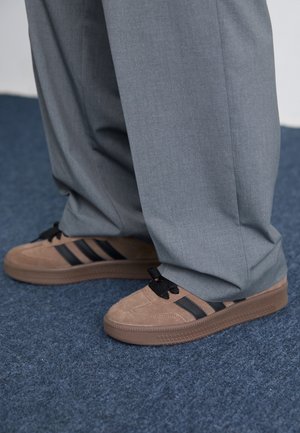 Personne portant un pantalon gris ample et des baskets en daim marron avec des rayures noires, debout sur un tapis bleu.