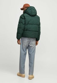 Grøn puffjakke med hætte, quiltet design og elastiske manchetter, kombineret med lyseblå denimbukser og beige sko.