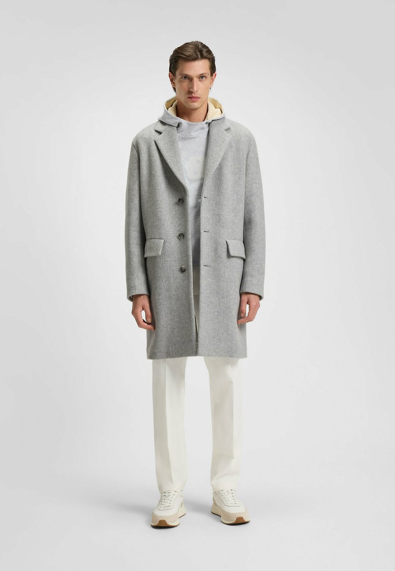 Cappotto di lana grigio con colletto a revers, design doppiopetto, due tasche frontali, indossato sopra un leggero hoodie, abbinato a pantaloni bianchi e sneakers.