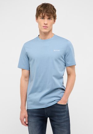 Junger Mann mit hellbraunen Haaren, der ein hellblaues Mustang-T-Shirt und dunkle Jeans trägt, steht mit einer Hand in der Tasche vor einem einfarbigen Hintergrund.