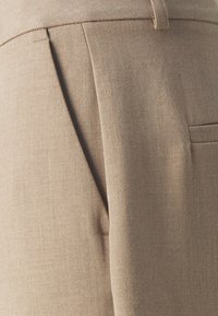 Béžové kalhoty na míru s kartáčovanou texturou, s předními sklady a zadní kapsou. Materiál je lehký a hladký.