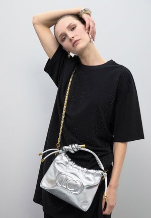 Frau trägt ein schwarzes übergroßes T-Shirt und goldene Armbänder, hält eine silberne Lederhandtasche mit goldener Kettenriemen und gerafftem oberen Teil.