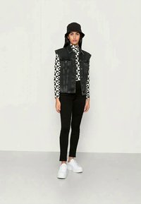 Colete de ganga preto sobre uma camisa xadrez preto e branco, combinado com calças pretas e ténis brancos. Complementado por um chapéu de balde preto.