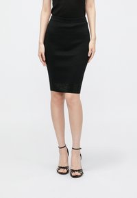 Čierna ceruzková sukňa po kolená z elastickej pleteniny, s hladkou textúrou a jemným detailom na leme. Kombinovaná s čiernymi sandálmi na remienkoch.