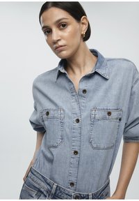 Light blue denim shirt with short sleeves, featuring two chest pockets, brown buttons, and a classic collar. Subtle denim texture noticeable.