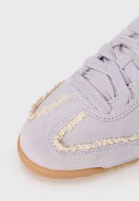 Gros plan sur la pointe d'une basket en daim violet clair avec une bordure en laine bouclée crème et une semelle en caoutchouc beige, équipée de lacets violets assortis.