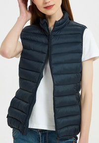 Le gilet matelassé marine présente un design matelassé horizontal, une fermeture éclair, un col montant et un tissu léger, superposé sur une chemise blanche.