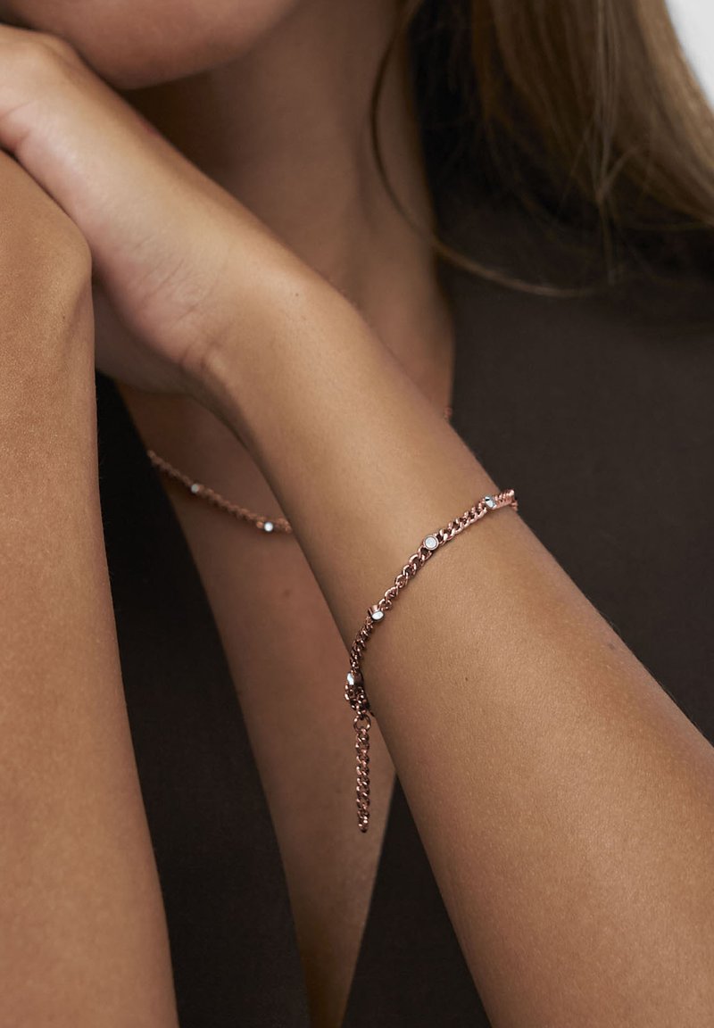 Roséguldskedja armband med små runda vita stenar som bärs på ett handled mot mörka kläder och ljus hudton.