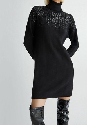 Mujer que lleva un vestido de punto negro con cuello alto y detalles de lentejuelas en el pecho, combinado con botas negras brillantes hasta la rodilla.