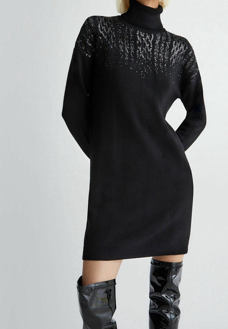 Femme portant une robe à col roulé en maille noire avec des détails en sequins sur la poitrine, assortie à des bottes noires brillantes montant jusqu'aux genoux.