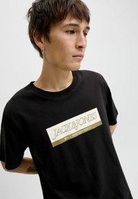 Sort bomulds T-shirt med et stort hvidt og guld logo-print, hvor teksten "JACK&JONES SINCE NINETEEN NINETY" fremhæves.