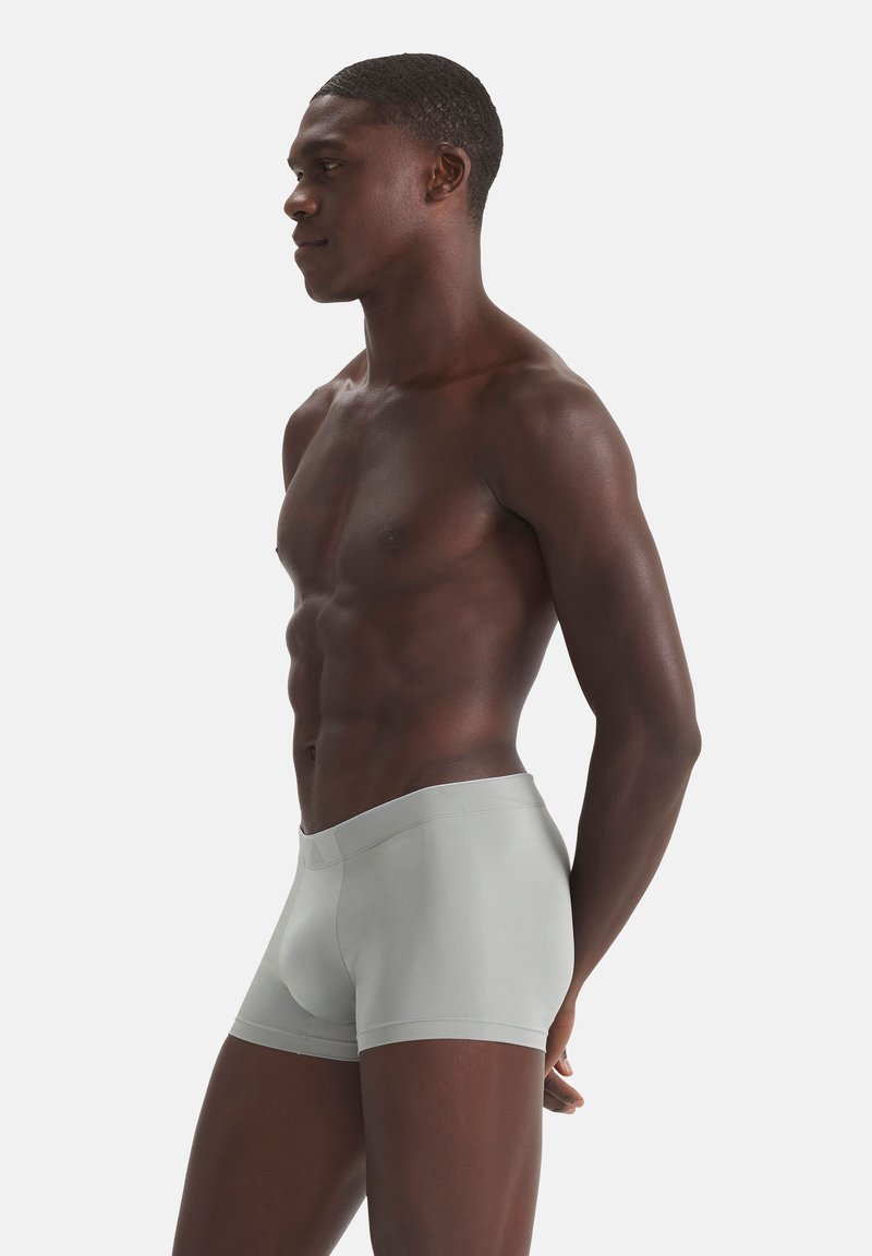 Modèle masculin debout de profil portant un boxer ajusté gris clair sur un fond blanc uni.