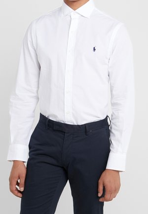 Man draagt een wit overhemd met knopen met een klein marineblauw polospelerlogo en donkerblauwe broek, staande tegen een effen achtergrond.