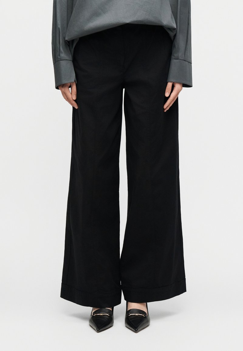 Άτομο που φοράει μαύρο παντελόνι με φαρδιά μπατζάκια, μαύρα αιχμηρά μοκασίνια με χρυσή αλυσίδα και χαλαρό γκρι μακρυμάνικο μπλουζάκι, στέκεται μπροστά σε λευκό φόντο.