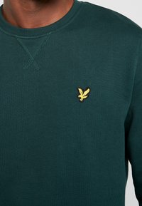 Dark green crewneck sweatshirt made of textured fabric. Features a small yellow and black embroidered bird logo on the chest.