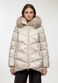 Giacca a vento beige lucida con motivo a chevron, dotata di cappuccio rifinito in pelliccia sintetica e tasche anteriori con zip. Tessuto liscio e taglio fino al ginocchio.