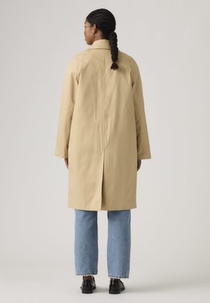 Une femme se tient de dos, portant un manteau beige aux genoux, un jean bleu clair et des chaussures noires, sur un fond de studio uni.
