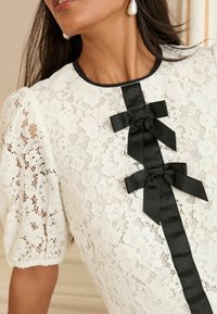 Maglietta bianca in pizzo con dettagli in nastro di raso nero, caratterizzata da un collo rotondo e maniche a sbuffo, con motivi floreali e tessuto strutturato.