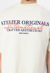 Lühikeste varrukatega kreemivärvi T-särk, mille seljal on mereväe ja oranži värvi tekst "ATELIER ORIGINALS" ja "CRAFTED AESTHETICS".