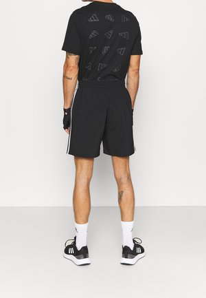 Man draagt zwarte Adidas shorts en T-shirt met witte strepen, zwarte sneakers, witte sokken en een zwarte vingervrije handschoen, staand met de rug naar voren.