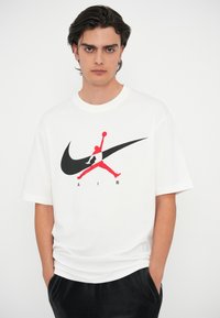 Balta medvilninė marškinėliai su dideliu juodu Nike swoosh logotipu ir raudonu Jumpman grafiku, žemiau esant tekstui "AIR". Atsitiktinis kirpimas, trumpi rankoviai.