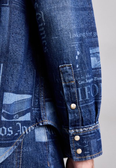 Denims krekls ar avīzes drukas dizainu zilu toņu gammā, ar pogām ap roku galiem un kontrastējošiem šuvju elementiem.
