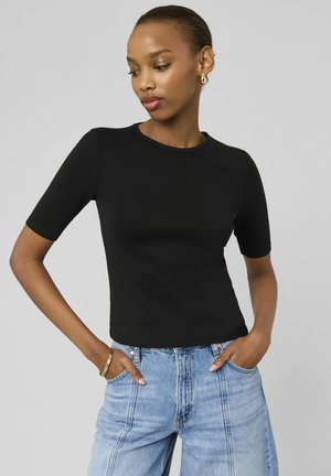 Femme portant un haut noir à manches courtes et un jean taille haute bleu clair, les mains dans les poches, debout devant un fond gris uni.