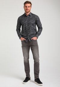 Camicia nera a fantasia con maniche lunghe, abbinata a jeans grigi e sneakers nere. Modello in piedi con le mani in tasca.