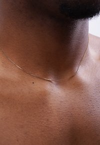 Dünner goldener Halsreif liegt auf der Haut und hebt die glatte, dunkle Hautstruktur hervor; subtiler Glanz und feine Verschlussdetails sind sichtbar.