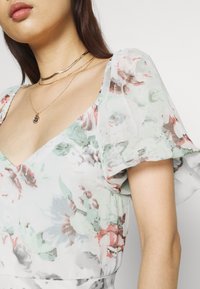 Robe florale légère en tissu doux ; présente des manches bouffantes, un décolleté en V et une palette de couleurs pastel de vert et de rose. Colliers superposés dorés.