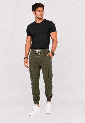 Ein Mann steht, trägt ein schwarzes T-Shirt, olivgrüne Cargohosen mit Kordelzug in der Taille, weiße Turnschuhe und eine Armbanduhr, schaut nach links.