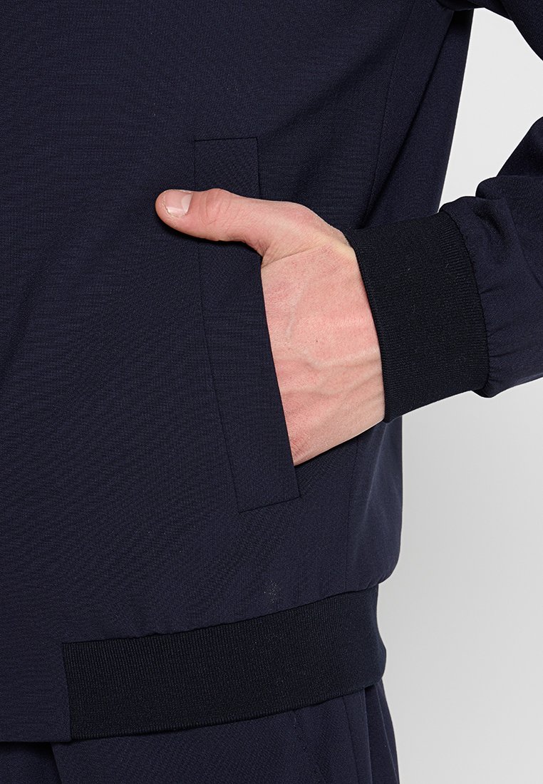 Marineblaues Jacket mit einer Seitentasche und strukturiertem Stoff, mit elastischem Bund und Ärmelbündchen für eine enganliegende Passform. Hand teilweise in der Tasche sichtbar.