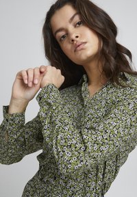 Camicia floreale verde con colletto, maniche lunghe e polsini con bottoni. Presenta un motivo di piccoli fiori bianchi e neri su uno sfondo verde.