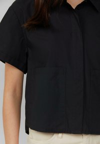 Chemise noire à manches courtes avec un devant boutonné, dotée de deux grandes poches plaquées et d'un ourlet droit. Le tissu semble lisse et léger.