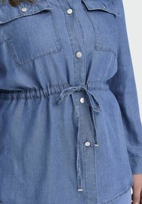 Denim srajca z dolgimi rokavi, dvema žepoma na prsih, pritisnimi gumbi, elastičnim vrvico za prilagajanje pasu in svetlo modro barvo. Gladka tekstura.