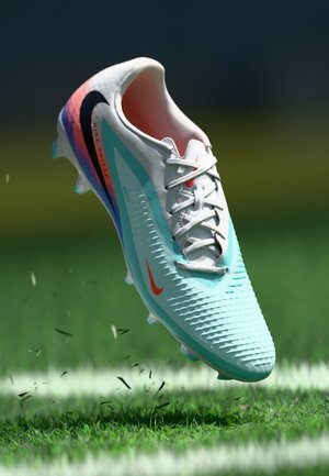 Nogometni čevlji Nike z lahkim modrim sintetičnim zgornjim delom, teksturiranimi valji, črnimi in oranžnimi poudarki ter belimi štikli, nad zeleno travo.