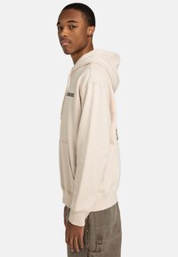 Haut à capuche beige avec cordon de serrage, poche kangourou et texte imprimé sur le devant. Texture douce, coupe décontractée, poignets et ourlet côtelés.