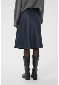 Jupe en satin marine avec un ourlet fluide, associée à des bottes de cowboy en cuir noir. Un pull gris tombe sur la taille, créant un look superposé.