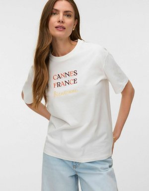 Kvinde med langt hår iført hvid T-shirt med teksten "Cannes France Bienvenue" og lyseblå jeans, stående mod en ensfarvet baggrund.
