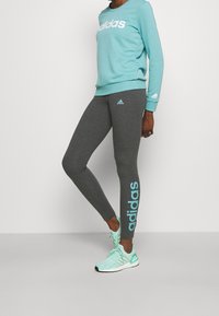 Turkos sweatshirt med vit "adidas"-logotyp, matchad med mörkgrå leggings med ljusblå "adidas"-text på sidan. Gröna träningsskor.