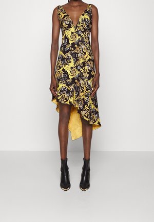 Woman wearing a black and yellow sleeveless asymmetrical dress with baroque patterns and black ankle boots with pointed gold toes.