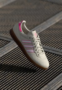 Sapatilha cinza claro com detalhes em rosa e três riscas, apresentando uma parte superior texturizada, solado de borracha e bico perfurado para respirabilidade.