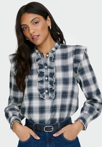 Rutet bluse i blå og hvid med rynket halsudskæring og ærmer. Har knaplukninger og bæres med et sort bælte samt blå denim bukser.