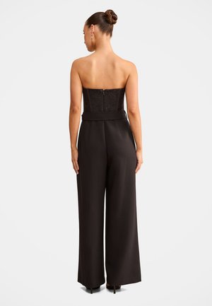 Femme aux cheveux attachés en chignon porte un top en dentelle noir sans bretelles et un pantalon noir à jambes larges, debout de dos sur un fond uni.