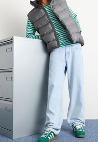 Vadderad grå väst över en grön och vit randig skjorta, ihop med ljusblå jeans och gröna sneakers med vita accenter, stående vid ett arkivskåp.