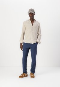 Camisa de lino color beige claro con frente abotonado, pantalones azul marino y zapatillas color tostado con un acento estampado. Una gorra gris completa el conjunto.