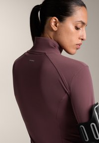 Burgundy träningslinne med hög krage, figurnära design, slät textur, med en logotyp på baksidan och ett svart armband med silverdetaljer.