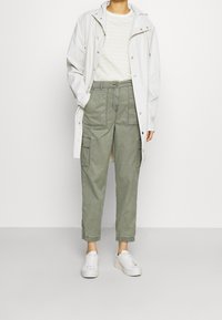 Lehká bílá nepromokavá bunda přes pruhovanou, světle béžovou košili. Olivově zelené cargo kalhoty s kapsami a bílé tenisky. Jednoduchý, ležérní outfit.