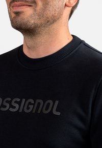 Felpa nera con colletto a coste e logo "Ossignol" testurizzato sul davanti. Il tessuto appare liscio con una finitura opaca.