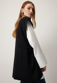 Hosszú vörös hajú nő ezüst fülbevalókkal, fehér hosszú ujjú blúzzal és fekete, oldalán megkötött ujjatlan mellénnyel.